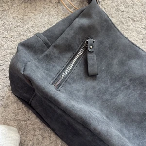 Mörkgrå mocka axelväska med dragkedja - Snygg mörkgrå axelväska i mocka med en praktisk dragkedja framtill. Väskan har en mjuk och stilren look, perfekt för att bära dina viktigaste grejer. Enkel design som passar till många outfits.