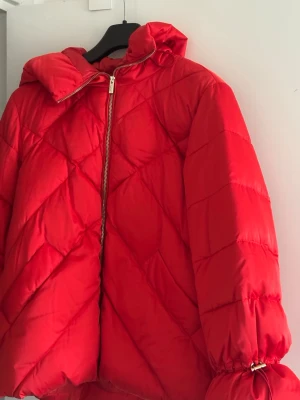 Röd pufferjacka från Marella  - Snygg röd pufferjacka från Marella sport med quiltat mönster och dragkedja i silver. Jackan har hög krage, luva och resår vid ärmsluten för extra komfort. Perfekt för kalla vinterdagar och ger en cool look till din outfit.