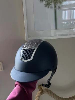 Mörkblå ridhjälm med glitterdetalj - Ordinarie pris 1 500kr. Snygg Marinblå ridhjälm från Crw med glittrig panel framtill och silvriga detaljer. Hjälmen har en klassisk skärm. Perfekt för dig som vill sticka ut i stallet och samtidigt ha bra skydd. Strl M 56-58, använda några gånger men aldrig tappade eller trillat av med. Har två stycket likadana, kolla sista bilden, har också en till annons på den andra. Pris kan absolut diskuteras, skriv om du har några frågor!