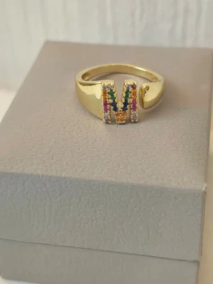 Guldfärgad ring med bokstaven M,  finns fler bokstäver  - Trendig guldfärgad ring med bokstaven M framtill, dekorerad med små färgglada stenar i regnbågens färger. Ringen har en bredare design och bokstaven är täckt av gnistrande stenar som ger en cool och personlig touch.     Justerbar I storlek och rostfritt stål 