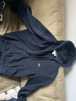 Mörkblå zip hoodie från Lacoste - Snygg mörkblå zip hoodie från Lacoste med dragkedja framtill och klassisk krokodil-logga på bröstet. Hoodien har huva med dragsko och två stora fickor framtill. Tillverkad i mjukt bomullsmaterial som är skönt att ha på sig. Jag säljer plagget eftersom det är lite litet på mig. Jag är 180 lång så jag skulle säga att det passar någon från 167-177 cm. Plagget har inga skador.