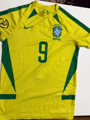 Brasilien Ronaldo 9 matchtröja Nike - Gul Brasilien fotbollströja från Nike med gröna detaljer, nummer 9 och Ronaldo på ryggen. Tröjan har CBF-emblemet och Nike-logga på bröstet. Tillverkad i Dri-FIT material som andas, perfekt för träning eller match.