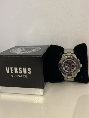 Versus Versace silverfärgad klocka - Stilren klocka från Versus Versace i silverfärgat metallarmband och svart urtavla med flera små urtavlor och tydliga markeringar. Klockan har en sportig design med detaljerade visare och Versus-logga på både urtavla och boett. OBS klockan har små skavanker och behöver ett nytt batteri! Extra länkar tillkommer, passar alla storlekar.