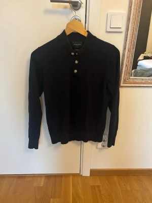  Mörkblå tröja med knappar från Grand Frank  - Stilren mörkblå långärmad tröja från Grand Frank med tre dekorativa knappar vid halsen. Tröjan har en klassisk rak passform och är tillverkad i mjukt material som passar perfekt till olika outfits.