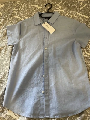 Ljusblå kortärmad skjorta Primark - Snygg ljusblå skjorta från Primark med klassisk krage och korta ärmar. Skjortan har knappar framtill och är gjord i mjuk bomull, perfekt för en clean och enkel stil. Passar dig som gillar minimalistiskt mode.