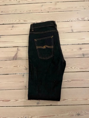 Mörkblå jeans från Nudie Jeans - Säljer ett par mörkblå jeans från Nudie Jeans med klassisk femficksmodell och kontrastsömmar. Jeansen har en rak passform och är tillverkade i kraftigt denimtyg. Snygg detalj med Nudie-loggan broderad på bakfickan.