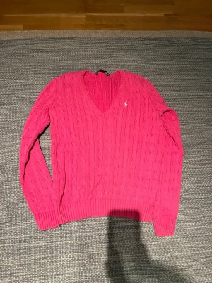 Rosa kabelstickad tröja från Polo Ralph Lauren - Säljer en snygg rosa kabelstickad tröja från Polo Ralph Lauren med V-ringning och klassisk vit logga broderad på bröstet. Tröjan har långa ärmar och ribbade muddar vid ärmslut och nederkant. Perfekt för dig som gillar stilrena och färgglada plagg.