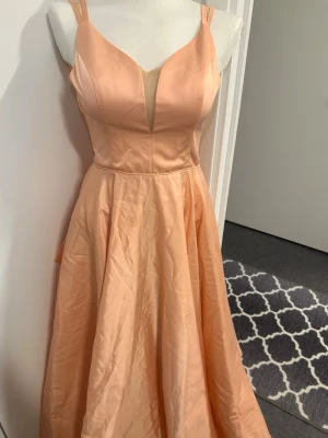Ljus persikofärgad långklänning - Elegant långklänning i ljus persikoton med djup v-ringning och breda axelband. Klänningen har en markerad midja och ett utsvängt kjolparti som ger en fin siluett. Tillverkad i ett glansigt material som ger en exklusiv känsla.