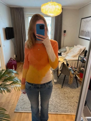 Stickad tröja i orange och gul - Säljer en snygg stickad tröja med färgskiftningar i orange, gul och vitt. Tröjan har lång ärm och är figurnära med rund halsringning. Materialet ser ut att vara mjukt och fluffigt, perfekt för att ge en cool och färgglad look.