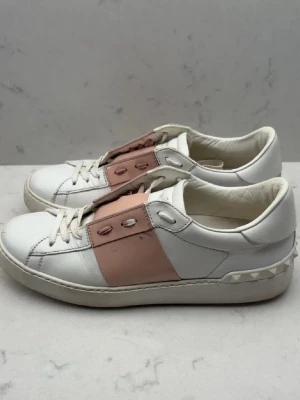 Valentino open  - Snygga sneakers från Valentino Garavani i vitt skinn med ljusrosa detalj över snörningen och ikoniska nitar på hälen. Klassisk rund tå och platt sula. Perfekta för dig som vill ha en stilren men ändå unik look. Äkta!!! Behöver säljas snabbt!!! Storlek 38 men sitter som 39! 