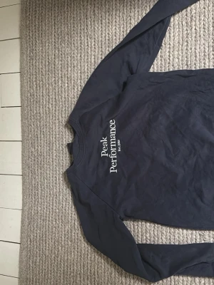 Mörkblå sweatshirt Peak Performance - Mörkblå sweatshirt från Peak Performance med vit logga på bröstet. Klassisk rund halsringning och långa ärmar. Tillverkad i mjukt material som passar perfekt till en avslappnad stil.