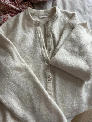 Vit kofta - Mysig vit kofta från Gina  med rund halsringning och pärlemorknappar framtill. Koftan är stickad och har lång ärm, perfekt för lager-på-lager. Enkel och stilren design som passar till det mesta.