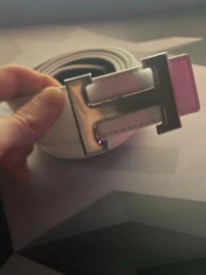 Vitt bälte med H-spänne - Snyggt vitt bälte med ett stort, silvrigt H-format spänne. Bältet är gjort i slätt läder och har stilrena sömmar längs kanterna. Perfekt accessoar för att lyfta din outfit med en modern touch.