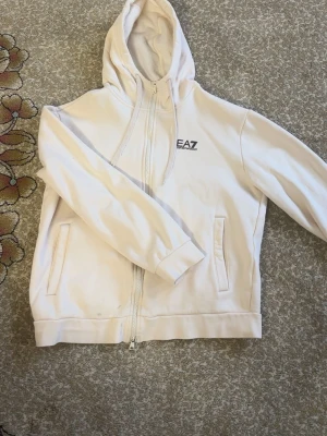 Beige zip up från EA7 Emporio Armani - Säljer en stilren beige hoodie från EA7 Emporio Armani med dragkedja framtill och två fickor. Hoodien har en klassisk huva och diskret logga på bröstet. Mjuk insida i fleece och ribbade muddar. Perfekt för en clean och avslappnad look.