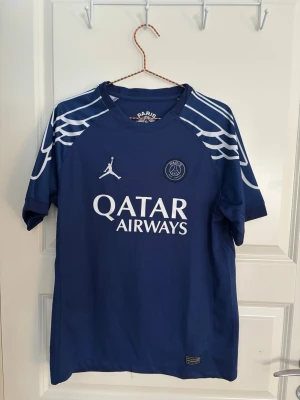 Blå PSG matchtröja Jordan - Säljer en blå PSG matchtröja från Jordan med vita detaljer och Qatar Airways-logga på bröstet. Tröjan har korta ärmar och coolt vingmönster på axlarna. Perfekt för fotbollsfans som vill representera sitt lag.