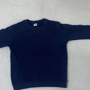 Mörkblå sweatshirt från C.P. Company - Snygg mörkblå sweatshirt från C.P. Company med rund halsringning och en unik detalj på ärmen – en liten ficka med dragkedja och den klassiska C.P. Company-linsen. Tillverkad i mjukt bomullsmaterial och har ribbade muddar vid ärmslut och nederkant. Storlek M men passar mer S.