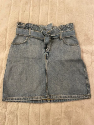 Blå jeanskjol med knyt från H&M - Säljer en snygg blå jeanskjol från H&M i storlek 36. Kjolen har ett brett knytband i midjan, två framfickor och en rak passform. Tillverkad i 100% bomull för en klassisk denimlook. Perfekt till sneakers eller boots! Ej bra pris,, skriv till mig