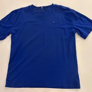 Blå t-shirt från Tommy Hilfiger - Snygg blå t-shirt från Tommy Hilfiger med liten broderad logga på bröstet. Klassisk passform och rund halsringning. Perfekt till jeans eller shorts för en clean och enkel look.
