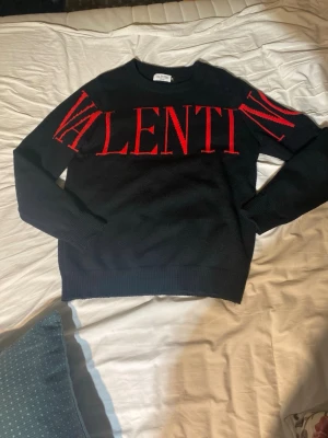 Svart stickad tröja från Valentino - Snygg svart stickad tröja från Valentino med stort rött VALENTINO-tryck över bröstet och axlarna. Tröjan har rund halsringning och ribbade muddar vid ärmslut och nederkant. Tillverkad i Italien och perfekt för dig som gillar exklusiv streetstyle.