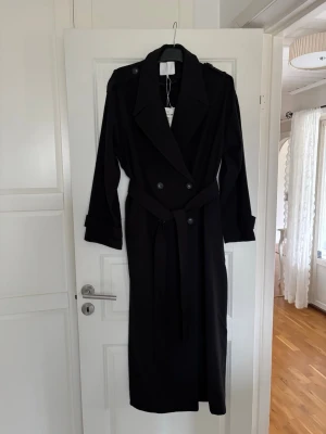 Svart lång trenchcoat med bälte - Stilren svart trenchcoat med dubbelknäppning, axelklaffar och bälte i midjan. Ärmarna har snygga detaljer med knappar och remmar. Klassisk krage och lång modell som ger en cool och tidlös look. Perfekt för dig som vill ha en trendig och mångsidig jacka.