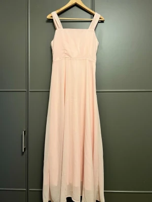 Ljusrosa långklänning med breda axelband - Elegant ljusrosa långklänning med breda axelband och rak skärning upptill. Klänningen har en mjukt draperad framsida och är tillverkad i ett tunt, flytande tyg som ger ett romantiskt intryck. Perfekt för sommarens speciella tillfällen.
