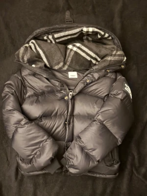 Svart buberry pufferjacka  - Snygg svart pufferjacka med stor vaddering och dragkedjor på fickorna. Jackan har en avtagbar huva med grått och vitt randigt innerfoder. Perfekt för kalla dagar och riktigt trendig look.