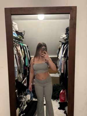 Grått bodyset med smala axelband - Säljer ett grått bodyset med smala svarta axelband. Setet består av en kort topp och matchande höga byxor med utsvängda ben. Materialet ser ut att vara stretchigt och bekvämt, perfekt för en trendig look.