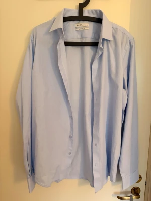 Ljusblå skjorta från S.C.W. - Klassisk ljusblå skjorta från S.C.W. i bomull med lång ärm och knappar framtill. Skjortan har en tidlös design med normal passform. Storleken passar M-L