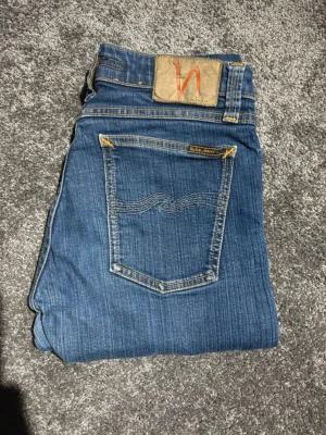 Nudie jeans 28/30 - Säljer ett par snygga blå slim fit jeans från Nudie i storlek 28/30. Klassisk modell med slim passform som sitter riktigt bra och passar till både vardag och mer uppklätt. Jeansen är i fint skick utan större slitningar – perfekta för dig som vill ha ett stilrent och hållbart plagg i garderoben.  Midja: 28 Längd: 30 Passform: Slim fit  Säljs då de tyvärr inte kommer till användning. Hör av dig vid frågor eller om du vill ha fler bilder! 😊