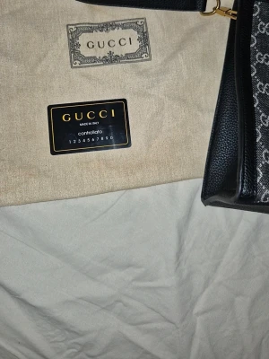 Svart handväska från Gucci med GG-mönster - Snygg svart handväska från Gucci med klassiskt GG-mönster i grått. Väskan har guldfärgade detaljer, bambuhandtag och en avtagbar axelrem. Tillverkad i canvas och läder, perfekt för dig som vill ha en lyxig och trendig accessoar.