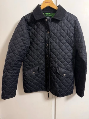 Svart quiltad jacka från Gant - Stilren svart quiltad jacka från Gant med klassisk krage i manchestertyg och tryckknappar framtill. Två stora fickor med lock och knapp, samt diskreta sömmar som ger en snygg passform. Perfekt för kyliga dagar och enkel att matcha.
