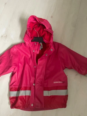 Rosa regnjacka från Didriksons - Snygg rosa regnjacka från Didriksons med avtagbar huva och reflexdetaljer nedtill. Jackan har fleecefoder på insidan för extra värme och tryckknappar framtill. Perfekt för regniga dagar och enkel att matcha med andra plagg.   Fleecefodrad