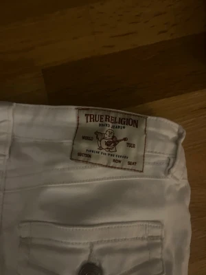 Vita jeans från True Religion - Säljer ett par vita jeans från True Religion med klassiska bakfickor och tydliga sömmar. Jeansen har raka ben och en snygg patch med logga bak i midjan. Perfekta för dig som vill ha en clean och stilren look.