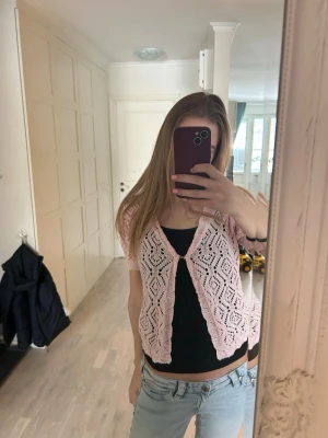 Ljusrosa virkad kofta med kort ärm - Söt ljusrosa kofta med korta ärmar och virkat mönster. Koftan har en öppen front med en liten knäppning upptill och är gjord i ett lätt material som passar perfekt över ett linne. Den har en avslappnad passform och fina detaljer i virkningen.