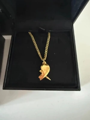 Guldigt halsband Maria Nilsdotter - Unikt halsband i guldton från Maria Nilsdotter med en cool berlock i form av ett ansikte. Kedjan är grov och smycket har en modern, edgy känsla. Perfekt för dig som vill sticka ut med ett statement piece i metall.