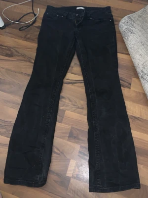 Svarta bootcut jeans med fickdetaljer - Säljer ett par svarta bootcut jeans med klassisk femficksdesign och snygga ficklock med knappar bak. Jeansen har normal midja och raka ben som går ut något nedtill. Perfekta till en avslappnad och trendig stil.