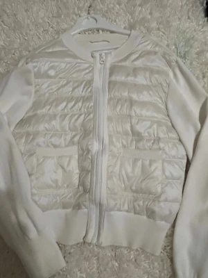 Vit quiltad cardiganjacka - Snygg vit cardiganjacka med quiltad framsida och två fickor. Jackan har dragkedja och ribbade muddar på ärmar och nederkant. Materialet är en mix av stickat tyg och glansigt quiltat tyg på framsidan, vilket ger en modern look.