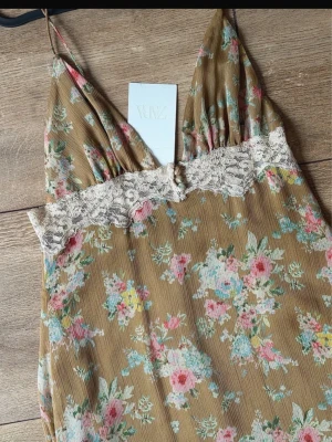 Blommig långklänning från Zara med prislapp - Säljer en romantisk långklänning från Zara med beige botten och blommigt mönster i rosa, blått och vitt. Klänningen har tunna axelband, spetsdetalj vid bysten och v-ringad hals. Perfekt för sommarens alla tillfällen!