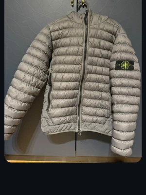 Grå dunjacka från Stone Island - Snygg grå dunjacka från Stone Island med quiltad design, dragkedja framtill och huva. Jackan är ej autentisk.har märkets ikoniska patch på vänster ärm och två sidofickor med dragkedja. Perfekt för kalla vinterdagar och riktigt trendig stil. Storlek M men passar S
