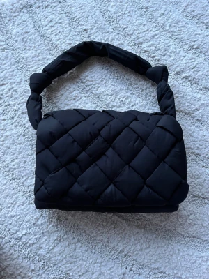 Svart quiltad handväska med knut - Trendig svart handväska med quiltat mönster och vadderad känsla. Väskan har en unik axelrem med knutdetaljer och är tillverkad i ett mjukt tygmaterial. Perfekt accessoar för att lyfta din outfit.