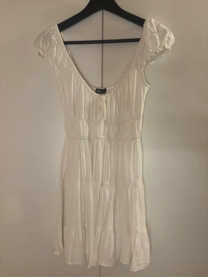 Vit kortklänning från New yorker - Söt vit kortklänning från New Yorker med puffärmar och smockdetaljer i midjan. Klänningen har en lätt rynkad struktur, rundad urringning med knyt och volangkant nedtill. Perfekt för varma dagar och sommarens alla tillfällen använt en gång och inte mer💕