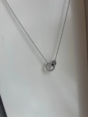 Silverhalsband med cirkelhänge - Snyggt halsband i silverfärg med ett tunt kedjeband och ett cirkelformat hänge dekorerat med små gnistrande stenar. En mindre, slät ring sitter i cirkeln.