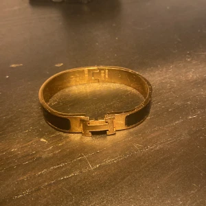 Guldigt armband med H-detalj - Snyggt armband i guldfärgad metall med en tydlig H-detalj framtill. Armbandet har en stilren och modern design med svarta emaljliknande partier på sidorna. Perfekt accessoar för att lyfta din outfit.