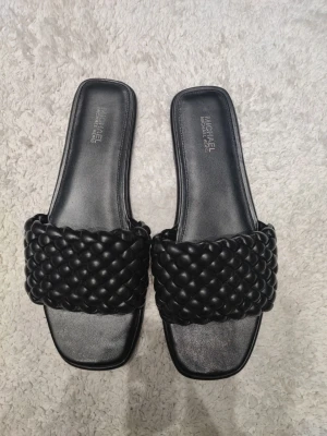 Svarta flätade sandaler Michael Kors - Stilrena svarta sandaler från Michael Kors med bred, flätad rem i vadderat konstläder. Sandalerna har fyrkantig tå och platt sula, perfekta för en trendig och avslappnad look. Passar till många olika outfits och ger en lyxig känsla.