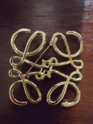 Guldfärgad brosch från Loewe - Säljer en elegant guldfärgad brosch från Loewe med deras ikoniska monogram i en snirklig, fyrkantig design. Broschen är tillverkad i metall och har en blank framsida samt detaljerad baksida. Perfekt accessoar för att lyfta din outfit.