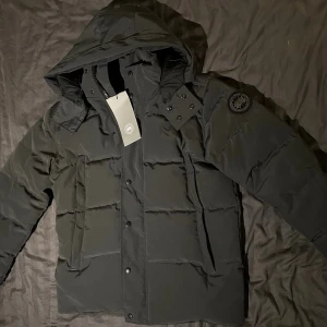 Canada Goose black label wynham parka - Aldrig använd med tagg kvar. Snygg svart dunjacka från Canada Goose med avtagbar huva och klassisk logotyp på ärmen. Jackan har en quiltad design, dragkedja och knappar framtill samt medföljande pälskrage. Perfekt för kalla vinterdagar. EJ ORGINAL!
