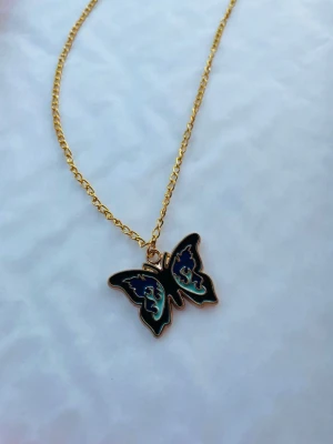 By prisförslag- Guldhalsband 45 cm med fjärilshänge - Snyggt halsband med guldfärgad kedja och ett svart och blått fjärilshänge med coola detaljer. Perfekt accessoar för att lyfta din outfit med en trendig touch. Kedjan är tunn och smidig, och fjärilen har en unik design med blå inslag.