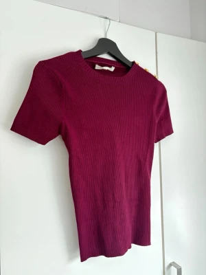 Vinröd ribbad topp med gulddetaljer - Snygg vinröd ribbad topp från Sfera  med korta ärmar och rund hals. Toppen har tre dekorativa guldfärgade knappar på ena axeln 