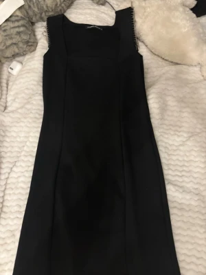 Svart kort klänning med fransar - Snygg svart kort klänning från Bar III med raka linjer och fyrkantig ringning. Ärmlös modell med coola silverfärgade fransdetaljer på axlarna som ger en unik touch. Perfekt för dig som vill sticka ut lite extra.