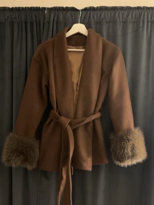 Brun kappa  - Snygg brun kappa med bred krage och knytskärp i midjan. Ärmsluten har fluffiga pälsdetaljer i brunt som ger en lyxig känsla. Perfekt för dig som vill sticka ut med en trendig och mysig look. Kappan är fodrad och har en avslappnad passform. Tyvärr för liten för mig nu men mycket bra skick fortfarande 